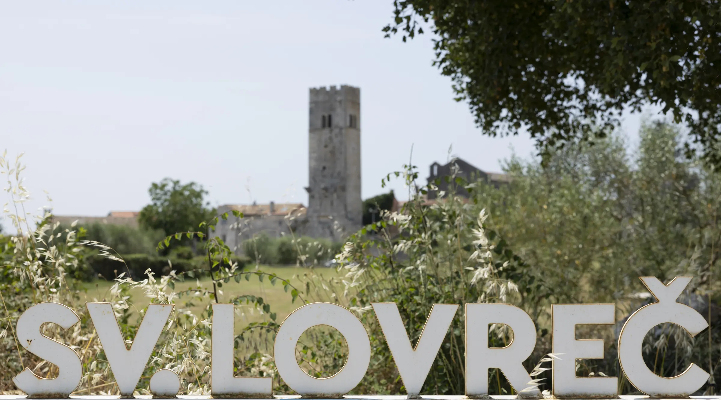 Weißer Schriftzug "SV. LOVREČ" im Vordergrund, historische Turmruine im Hintergrund, umgeben von grünen Olivenbäumen und Sträuchern.