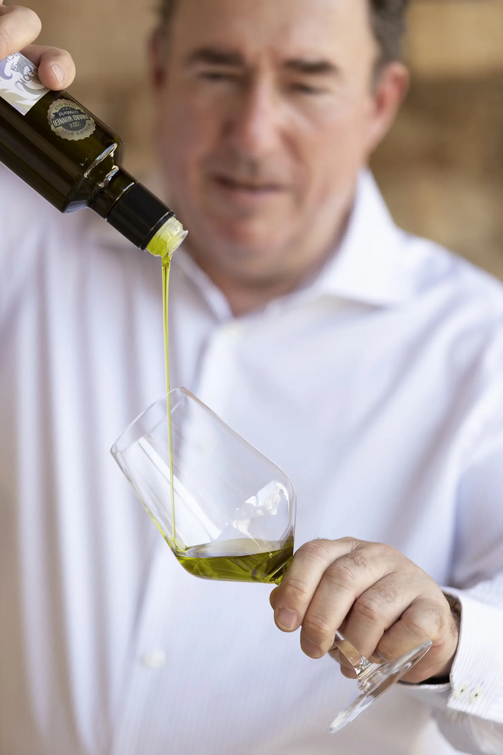 Ein Mann in einem weißen Hemd schenkt grünes Olivenöl in ein Glas, das bei Tageslicht auf einem blauen Hintergrund schimmert.