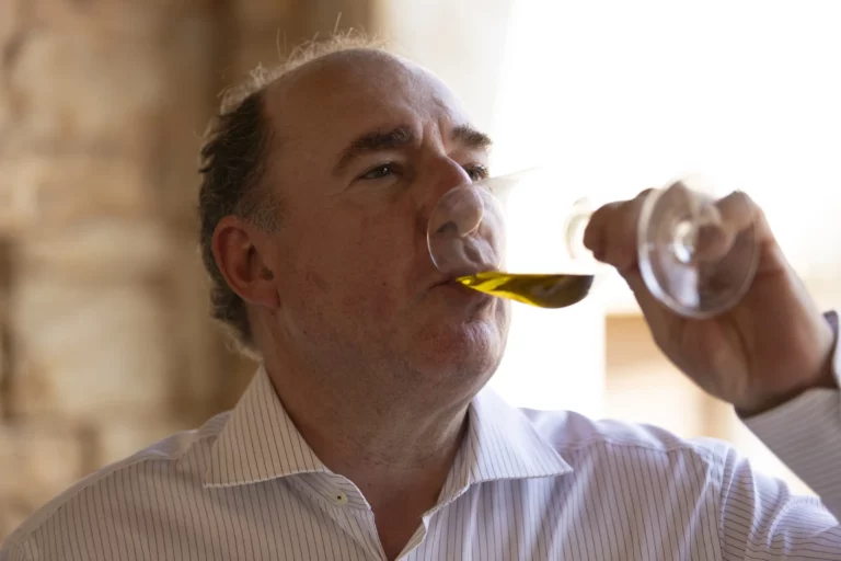 Mann in gestreifter Hemd hält ein Glas mit grünlich-gelbem Olivenöl und kostet es, im Hintergrund helles, rustikales Ambiente.