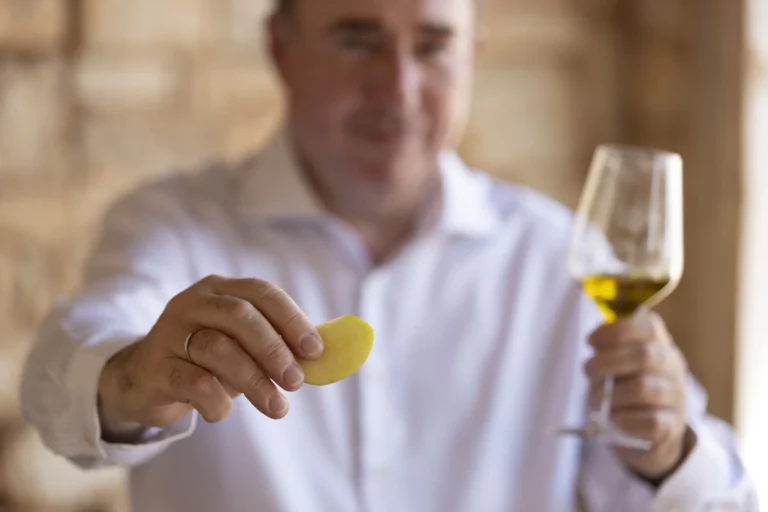 Rudolf Nemetschke in weißem Hemd präsentiert eine Apfelscheibe bei einer Olivenöl-Degustation und rechts hält er ein Glas mit goldenem Olivenöl.