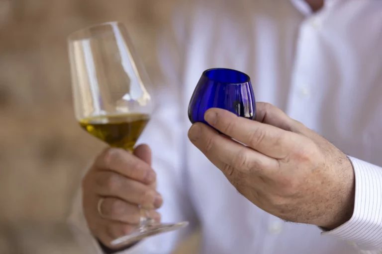 Ein Mann in einem weißen Hemd hält ein Glas mit grünlichem Olivenöl in der einen Hand und ein blaues Glas in der anderen.