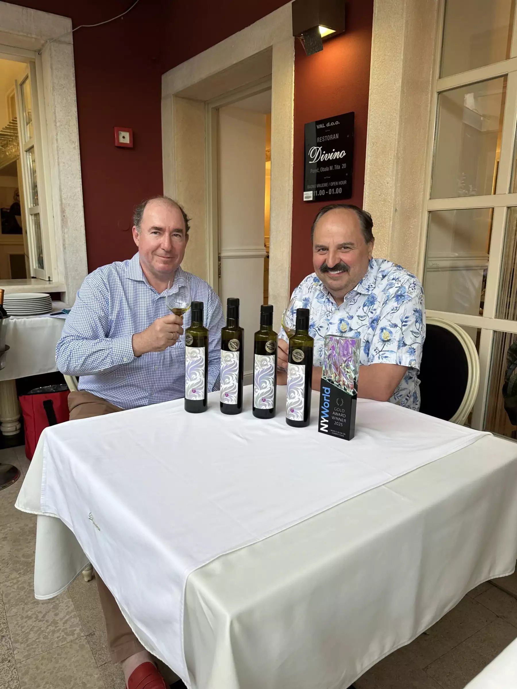 Rudolf Nemetschke sitzt mit Promikoch Johann Lafer in einem Gastgarten im Restaurant Divino. Vor Ihnen stehen 4 Avistria Olivenölflaschen zur Verkostung bereit, auch dabei der Gold Award der NY World.