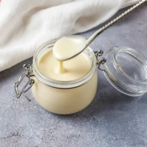 Auf einer steinernen Arbeitsplatte sieht man einen Löffel der in ein geöffnetes Einmachglas, gefüllt Goldene Mayonnaise, eintaucht und im Hintergrund liegt ein Geschirrtuch