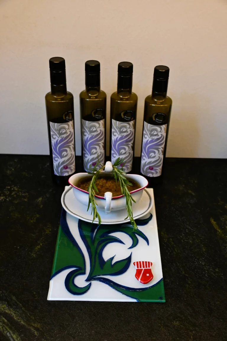 Im Hintergrund sieht man 4 Avistria Olivenölflaschen, davor steht eine Keramikschale gefüllt mit Preporuka Maslinara auf einer Glasfliese im Avistria Design