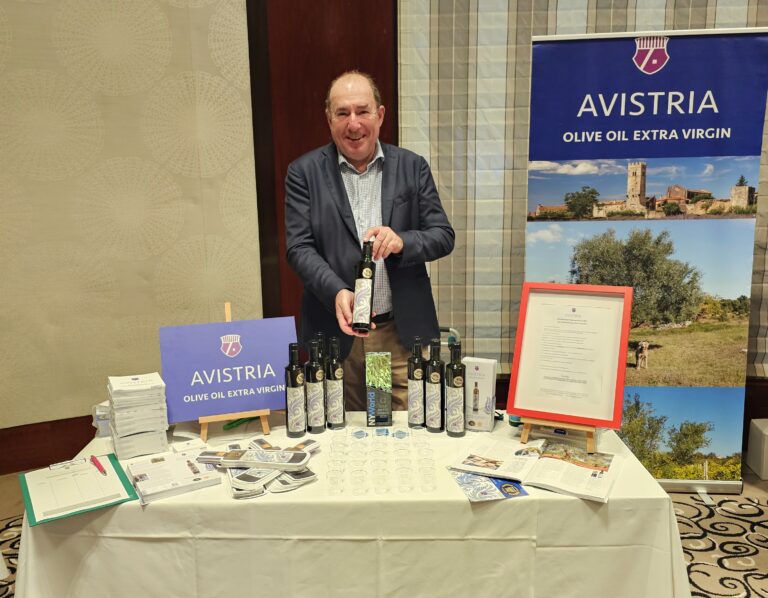 Rudolf Nemetschke präsentiert eine Flasche Olivenöl vor einem Stand mit dem Namen "Avistria Leccino", umgeben von Informationsmaterial und Ausstellungsbildern.